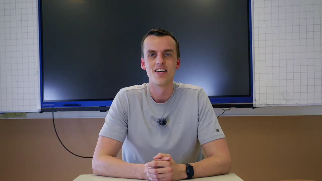 Menno van Math with Menno zit aan een tafel voor een digibord en lacht naar de camera